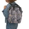 Smiling person in a denim jacket and jeans carries the Camo Diaper Backpack With Hunting Concealment Camouflage Sitka Optifade Open Country over one shoulder.