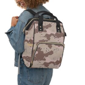 A smiling woman with curly hair in a blue denim jacket and jeans carries the Camo Diaper Backpack With Military Concealment Camouflage Desert Storm Chocolate Chip over her shoulder.